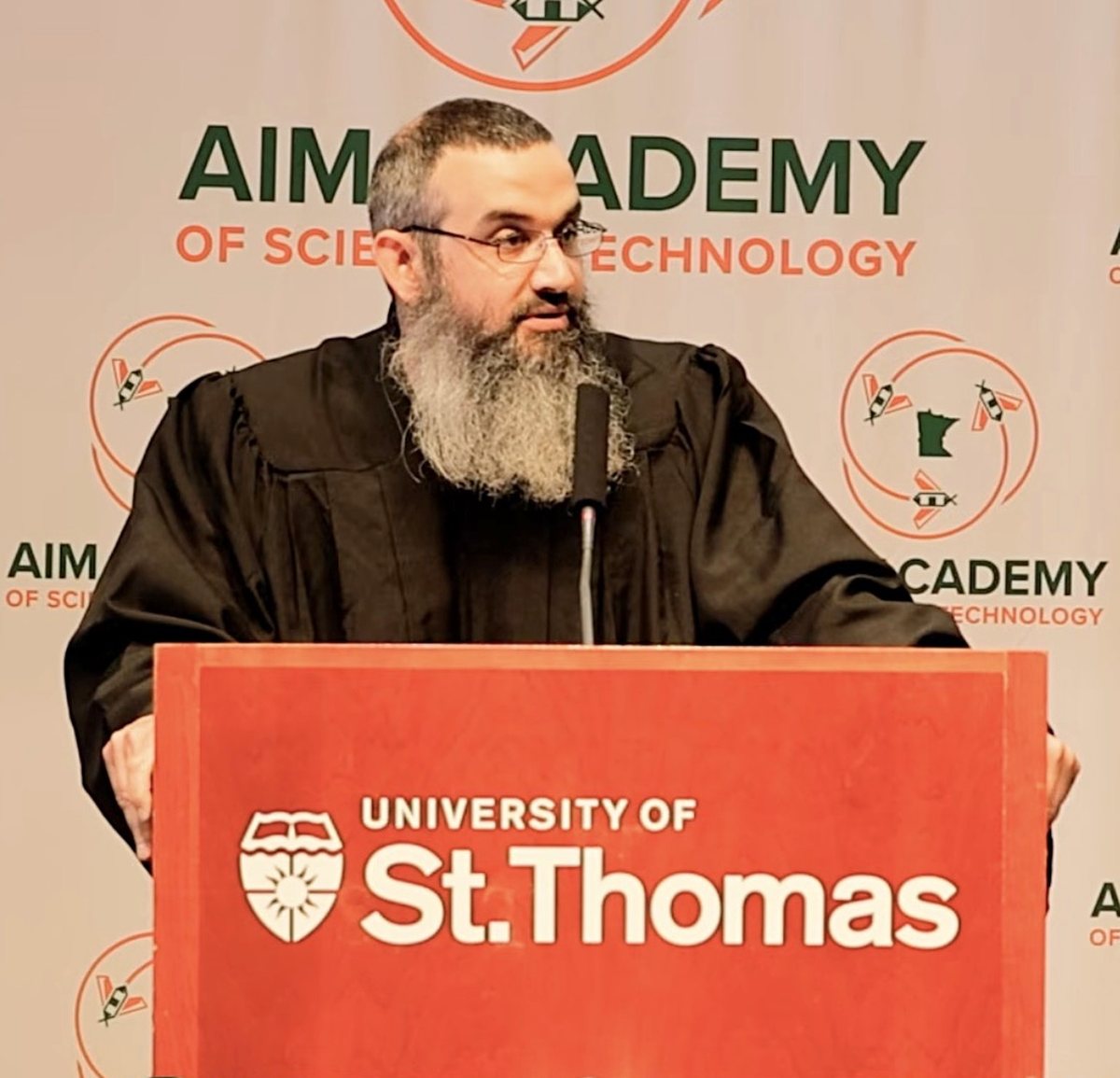 Michael Abraham delivering a graduation address at a University of St. Thomas podium, AIM Academy backdrop